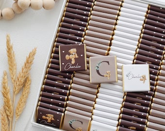 May include: A box of rectangular chocolate bars wrapped in dark brown, light brown, and white paper. Some wrappers feature a teddy bear illustration, the name Charlie, or the letter C with floral accents. Small white hearts decorate the dark bars.