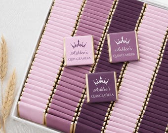 Könnte beinhalten: Eine Sammlung quadratischer Schokoladentafeln, eingewickelt in hellrosa, malvenfarbene und dunkelviolette Folie. Zwei Tafeln zeigen eine weiße Kronengrafik und den aufgedruckten Text Ashlee's QUINCEAÑERA. Die Tafeln sind in einem Raster in einer Box angeordnet.