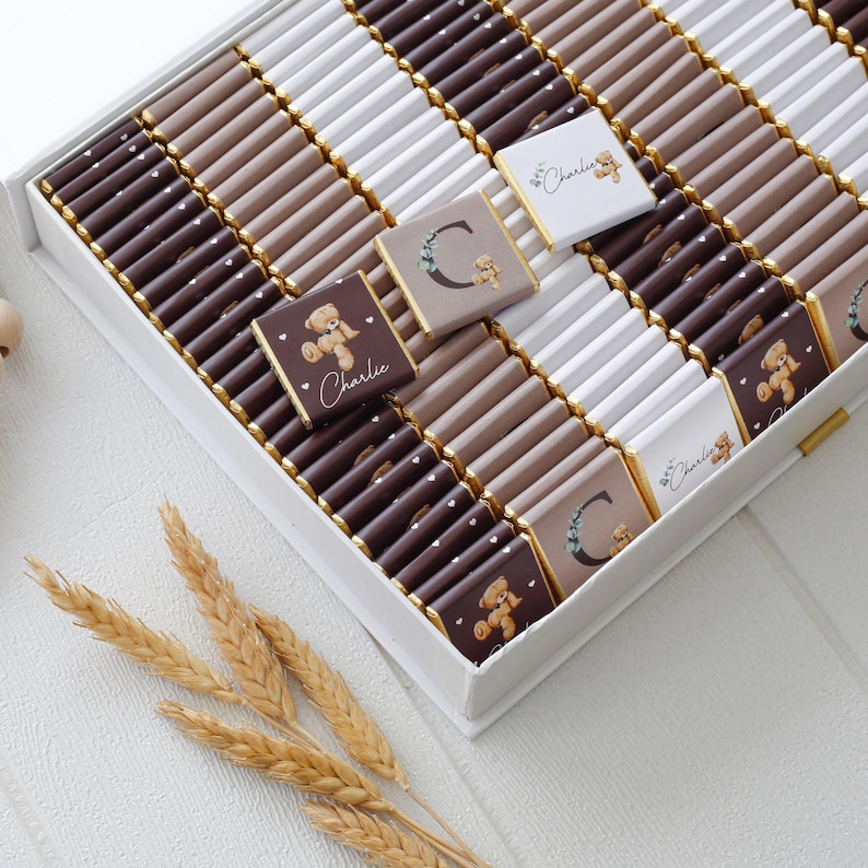 May include: A white box filled with individually wrapped chocolates in shades of brown, beige, and white. Some chocolates have a teddy bear graphic and the name "Charlie" printed on them. The box is decorated with gold trim.