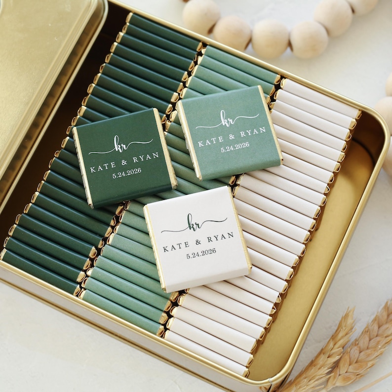 Pu&ograve; includere: Una scatola di metallo dorato riempita con file di piccole barrette di cioccolato rettangolari avvolte in carta verde scuro, verde salvia e bianca. Tre barrette sono poste sopra, stampate con le iniziali kr, KATE & RYAN e la data 5.24.2026.