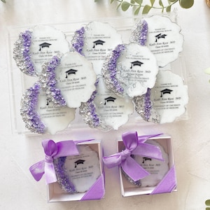 May include: Purple and silver graduation party favors with a white background. The favors are round and have a white background with the text "Kadi-Ann Rose MD Class of 2023 University of Cincinnati College of Medicine" printed on them. The favors are packaged in small white boxes with purple ribbon.