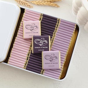 Pu&ograve; includere: Una scatola di metallo bianca riempita con file di piccole barrette di cioccolato avvolte in carta rosa chiaro, viola scuro e lavanda. Tre barrette presentano un grafico di due anelli intrecciati, il testo SARAH & JACOB e la data 05.07.2026.