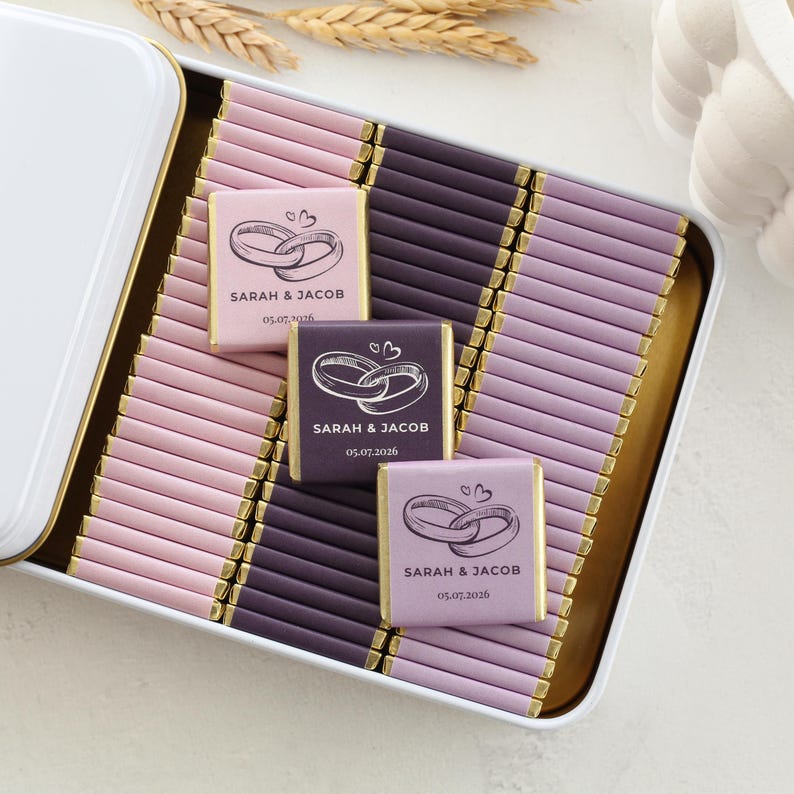 Pu&ograve; includere: Una scatola di metallo bianca riempita con file di piccole barrette di cioccolato avvolte in carta rosa chiaro, viola scuro e lavanda. Tre barrette presentano un grafico di due anelli intrecciati, il testo SARAH & JACOB e la data 05.07.2026.