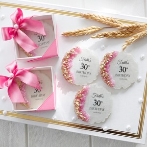 May include: A white tray holds two gift boxes with pink bows and three decorative coasters. The coasters have a white base, pink and gold accents, and the text "Faith's 30th Birthday 12.26.2025".