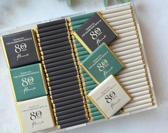May include: A clear box filled with small chocolate bars wrapped in gold and green paper. The wrappers have the text "Thank you for celebrating with us 80 Konna" printed on them.