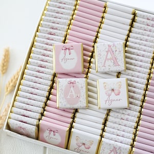 May include: A box of individually wrapped chocolates with pink and white floral designs. Some chocolates feature a pink bow, a butterfly, or the name "Aynur". The chocolates are arranged in rows, creating a visually appealing presentation.