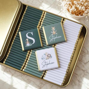 May include: A gold-rimmed tin filled with individually wrapped chocolates. Three chocolates are visible, each with a different design: a large white letter "S" with animal illustrations, a giraffe with the name "Stephen", and a collection of animals with the name "Stephen".