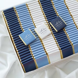 Pu&ograve; includere: Una grande collezione di barrette di cioccolato rettangolari avvolte in carta blu navy, azzurro e bianca con bordi in foglia d'oro. Tre barrette sono posizionate sopra, ognuna con un logo circolare con il testo With love Kasey & Marc.