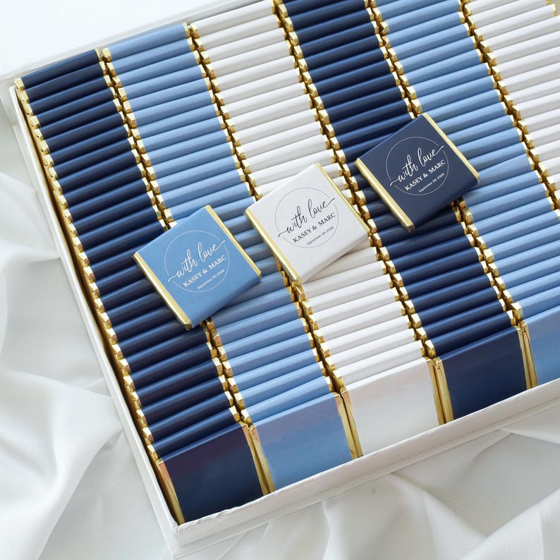 Pu&ograve; includere: Una grande collezione di barrette di cioccolato rettangolari avvolte in carta blu navy, azzurro e bianca con bordi in foglia d'oro. Tre barrette sono posizionate sopra, ognuna con un logo circolare con il testo With love Kasey & Marc.