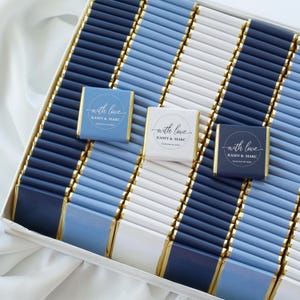 Pu&ograve; includere: Una collezione di barrette di cioccolato rettangolari avvolte in carta stagnola blu navy, azzurro e bianca. Tre barrette presentano un'etichetta circolare con il testo with love, Kasey & Marc e September 28, 2024, disposte in una scatola.
