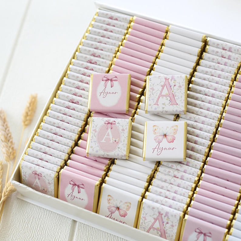 May include: A box of chocolates with pink and white wrappers. Some chocolates have the name "Aynur" and a butterfly or the letter "A" printed on them. The chocolates are arranged in rows inside a white box.
