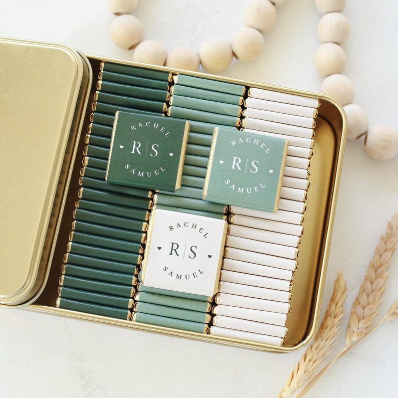 Pu&ograve; includere: Una scatola di metallo dorato riempita con file di piccole barrette di cioccolato avvolte in carta verde scuro, verde salvia e bianca. Tre barrette presentano il testo stampato Rachel, Samuel e le iniziali RS con piccoli cuori.