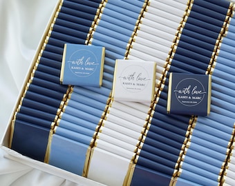 May include: A collection of rectangular chocolate bars wrapped in navy blue, light blue, and white foil. Three bars feature a circular label with the text with love, Kasey & Marc, and September 28, 2024, arranged in a grid pattern within a box.