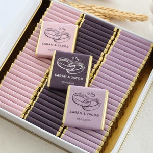 Pu&ograve; includere: Una scatola bianca riempita con file di piccole barrette di cioccolato rettangolari avvolte in carta rosa chiaro, viola scuro e lavanda. Ogni barretta presenta un grafico di due anelli intrecciati, due piccoli cuori e il testo Sarah & Jacob 05.07.2026.