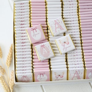 May include: A box of individually wrapped chocolates, primarily pink and white. Some chocolates feature a pink letter "A" or a butterfly design. Others have a bow or floral pattern, with the name "Aynur" printed on them. The chocolates are arranged in rows.