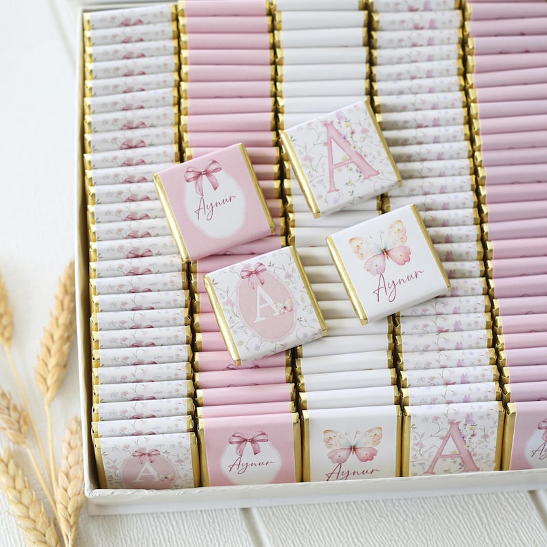 May include: A box of individually wrapped chocolates, primarily pink and white. Some chocolates feature a pink letter "A" or a butterfly design. Others have a bow or floral pattern, with the name "Aynur" printed on them. The chocolates are arranged in rows.