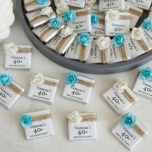 Op de afbeelding: Witte chocoladerepen met de tekst "Vanessa's 40th BIRTHDAY PARTY", versierd met touw en blauwe en witte papieren bloemen. De repen zijn gerangschikt op een grijze schaal.