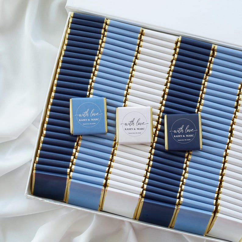 Pu&ograve; includere: Una grande scatola bianca riempita con file di piccole barrette di cioccolato rettangolari avvolte in carta stagnola blu navy, azzurro e bianca. Tre barrette presentano un'etichetta circolare con il testo "with love KASEY & MARC" e una data stampata in bianco.