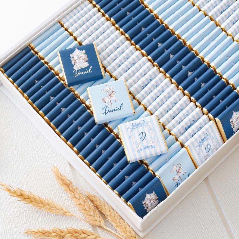 May include: A white box filled with individually wrapped chocolates. The chocolates are in shades of blue and white, some with a striped pattern. Some chocolates have the name "Daniel" or the letter "D" printed on them.