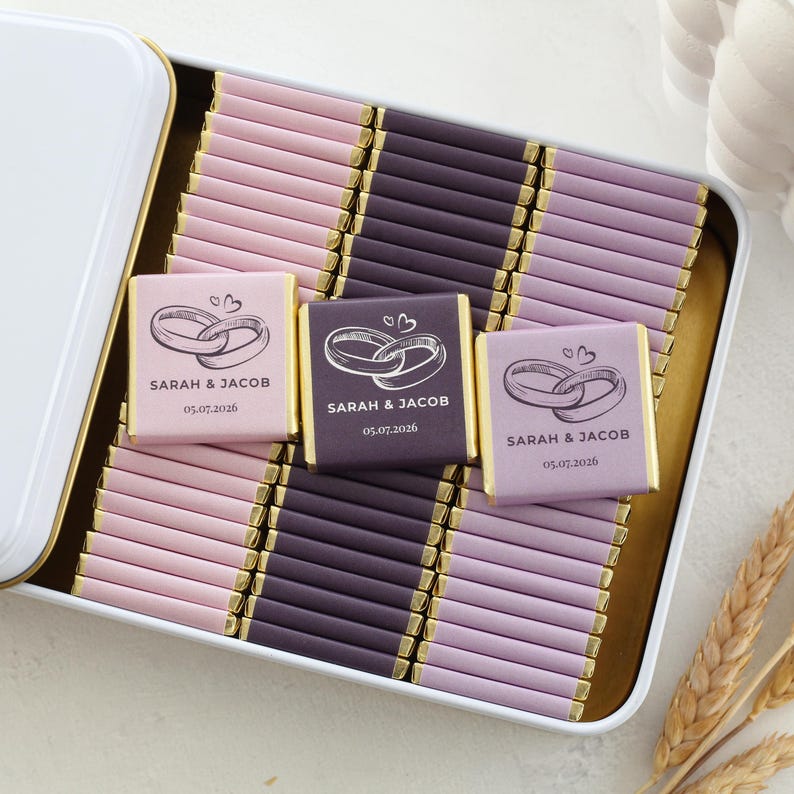 Pu&ograve; includere: Una scatola di metallo bianca riempita con file di piccole barrette di cioccolato avvolte in carta rosa chiaro, viola scuro e lavanda. Tre barrette presentano una grafica stampata di due anelli intrecciati, i nomi Sarah & Jacob e la data 05.07.2026.