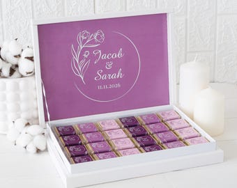 Può includere: Scatola regalo bianca aperta, che rivela cioccolatini. L'interno della scatola è viola con testo bianco: "Jacob & Sarah" e la data "11.11.2026". I cioccolatini sono avvolti in tonalità viola e rosa, disposti ordinatamente.