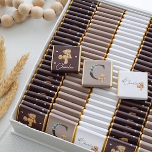 May include: A box of chocolates with a variety of colors and designs. Some chocolates have a teddy bear graphic and the name "Charlie" printed on them. Other chocolates have the letter "C" and a teddy bear graphic. The chocolates are arranged in rows.