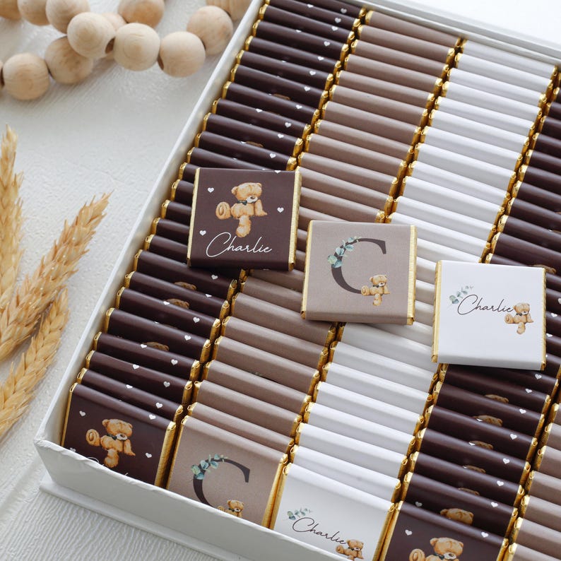 May include: A box of chocolates with a variety of colors and designs. Some chocolates have a teddy bear graphic and the name "Charlie" printed on them. Other chocolates have the letter "C" and a teddy bear graphic. The chocolates are arranged in rows.