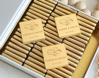 Könnte beinhalten: Eine weiße Dose gefüllt mit goldverpackten Schokoriegeln. Drei goldene Quadrate mit den Namen "SARAH & JACOB" und dem Datum "05.07.2026" sind sichtbar. Jedes Quadrat zeigt ein eingraviertes Design von zwei ineinander verschlungenen Ringen.