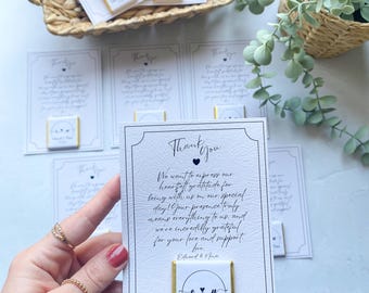 Può includere: Un biglietto di ringraziamento bianco con testo nero e un cuore nero. Il biglietto dice "Grazie" e include un messaggio che esprime gratitudine per aver partecipato a un matrimonio. Una piccola barretta di cioccolato bianco con foglia d'oro e le iniziali "E & N" è attaccata al biglietto.