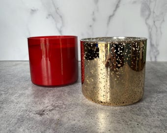 May include: Two cylindrical candle vessels are displayed. One vessel is a solid, opaque red glass, while the other is a metallic gold glass featuring a speckled, mercury-style finish that creates a reflective, textured appearance on the exterior surface.