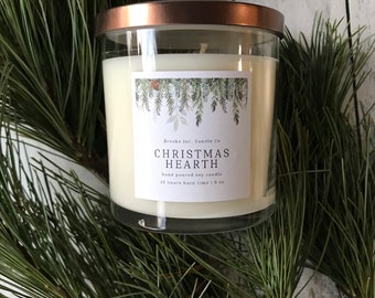 May include: A clear glass candle jar with a copper-colored lid, labeled "CHRISTMAS HEARTH." The label features a botanical design. The candle is surrounded by green pine branches. The candle is hand-poured soy and has a 45-hour burn time.