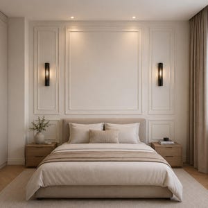Pode incluir: Um quarto com uma cama estofada em bege, almofadas a condizer e uma manta, ladeada por duas mesas de cabeceira de madeira. A parede atr&aacute;s da cama tem molduras brancas e dois candeeiros de parede verticais pretos com luz quente.