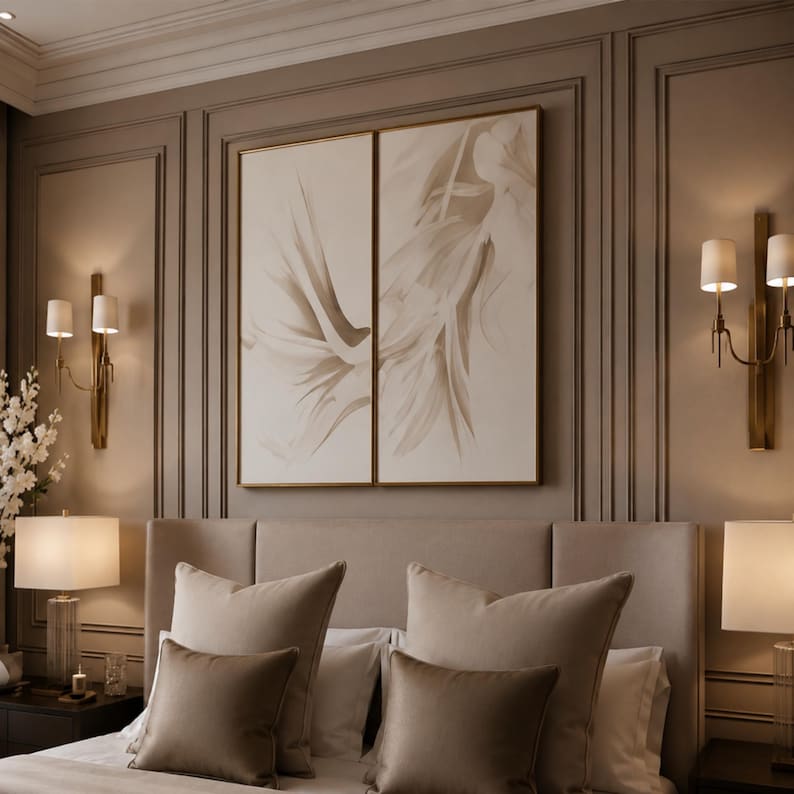 May include: A diptych wall art set featuring abstract beige and white brushstrokes in thin gold frames, mounted above a beige upholstered headboard with matching beige pillows, flanked by two table lamps and two wall-mounted sconces.
