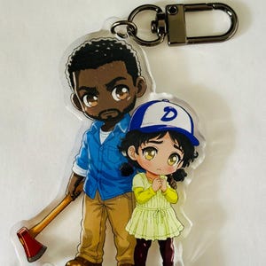 May include: An acrylic keychain featuring a cartoon illustration of a man with a beard holding an axe and a child wearing a blue and white baseball cap with a letter D. The keychain includes a dark metal lobster clasp attachment at the top.