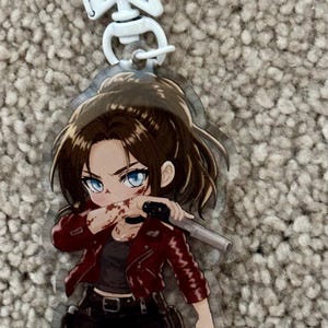 May include: An acrylic keychain featuring a chibi-style character with brown hair in a ponytail, wearing a red jacket, black top, and black pants. The character has blue eyes, blood splatters on their face and arm, and holds a silver handgun.