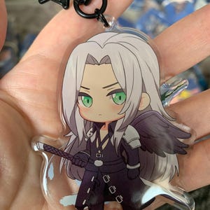 May include: A clear acrylic keychain with a chibi-style illustration of a character with long silver hair and green eyes. The character wears a black outfit with a single black wing and holds a sword. The keychain has a black star-shaped clasp.