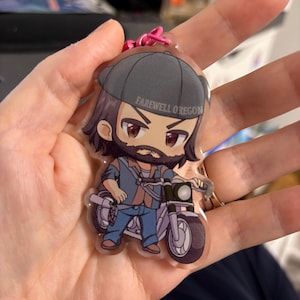 Könnte beinhalten: Ein klarer Acryl-Schlüsselanhänger mit einer Comicfigur mit Bart, die eine graue Kappe und blaue Jeans trägt und Motorrad fährt. Der Schlüsselanhänger hat einen rosa Verschluss und den Text "FAREWELL OREGON".