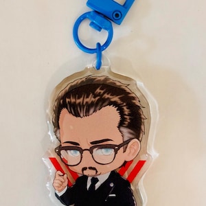 Könnte beinhalten: Ein Acryl-Schlüsselanhänger mit einer Cartoon-Illustration einer Person mit dunklem Haar, Ziegenbart und Brille, bekleidet mit einem schwarzen Anzug und Krawatte. Die Figur steht vor zwei roten V-förmigen Linien. Ein blauer Kunststoff-Karabiner ist befestigt.
