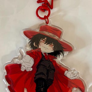 May include: An acrylic keychain featuring a stylized anime character with black hair and red eyes, wearing a red hat, a long red coat, and a black suit. The character has white gloves and is reaching forward. The keychain includes a red star-shaped clasp.
