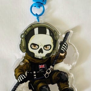 May include: An acrylic keyring featuring a cartoon character wearing a skull mask, olive green tactical gear, and a headset. The character has a black vest with a Union Jack flag and the text SAS, and is holding a black rope with a blue plastic clip.