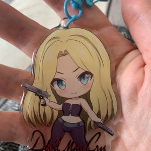 May include: A clear acrylic keychain with a cartoon image of a woman with blonde hair and blue eyes, holding two guns. The keychain has a blue star-shaped clasp and the words "Devil May Cry" in a script font.