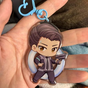 Könnte beinhalten: Ein klarer Acryl-Schlüsselanhänger mit einem Charakter im Chibi-Stil mit braunen Haaren, der einen Anzug trägt und eine Waffe hält. Der Schlüsselanhänger hat einen hellblauen herzförmigen Verschluss.