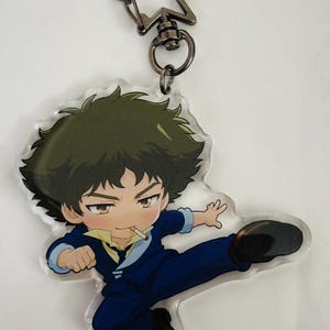 Peut inclure: Un porte-clés en acrylique représentant un personnage de dessin animé aux cheveux foncés en bataille, vêtu d'un costume bleu avec une chemise et une cravate jaunes. Le personnage donne un coup de pied, une cigarette à la bouche.