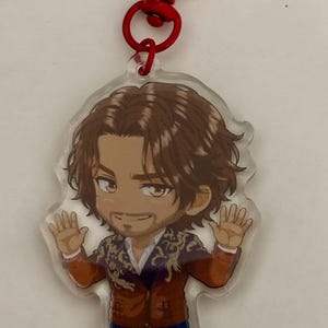 May include: A clear acrylic keychain featuring a cartoon illustration of a person with brown hair and a beard, wearing a brown jacket with gold embroidery and blue pants. The figure has both hands raised and is attached to a red heart-shaped clasp.