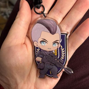 Puede incluir: Un llavero acrílico transparente con un personaje de dibujos animados con cabello púrpura y ojos azules, sosteniendo un rifle. El llavero tiene un cierre negro y un emblema en forma de escudo con un diseño dorado y blanco.