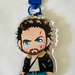May include: An acrylic keychain featuring a chibi-style character with dark brown hair, blue eyes, and a beard. The character wears a black jacket with a tan collar, a white shirt, and blue jeans, holding a silver revolver with red marks on the face.