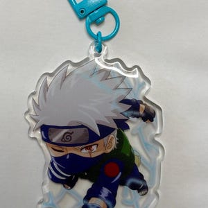 May include: A clear acrylic keyring featuring a cartoon character with spiky silver hair, a blue headband with a metal plate, and a dark blue face mask. The character wears a green vest and blue outfit, with light blue lightning bolt patterns.