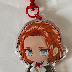 以下が含まれることがあります： オレンジ色の髪と青い目をしたちびキャラのアクリルキーホルダー。キャラクターは白いファーの襟が付いたダークブラウンのジャケット、白いシャツ、赤いネッカチーフ、茶色のズボンを着用しています。赤いハート型の留め具が付いています。