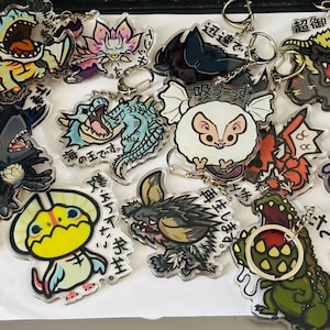 May include: A collection of clear acrylic keychains featuring colorful, stylized illustrations of various fantasy creatures and characters, each with Japanese text printed on the surface and attached to a metal split ring for hanging.