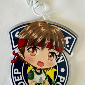 May include: A clear acrylic keychain featuring a chibi-style character with brown hair, a red headband, and a green and white uniform. The character is positioned over a circular dark blue emblem with white text reading DEP and POL.